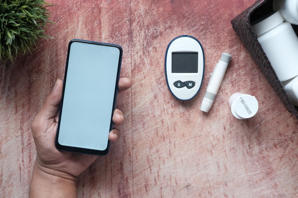 Hand holding a smartphone next to a glucometer and health supplies on a wooden surface